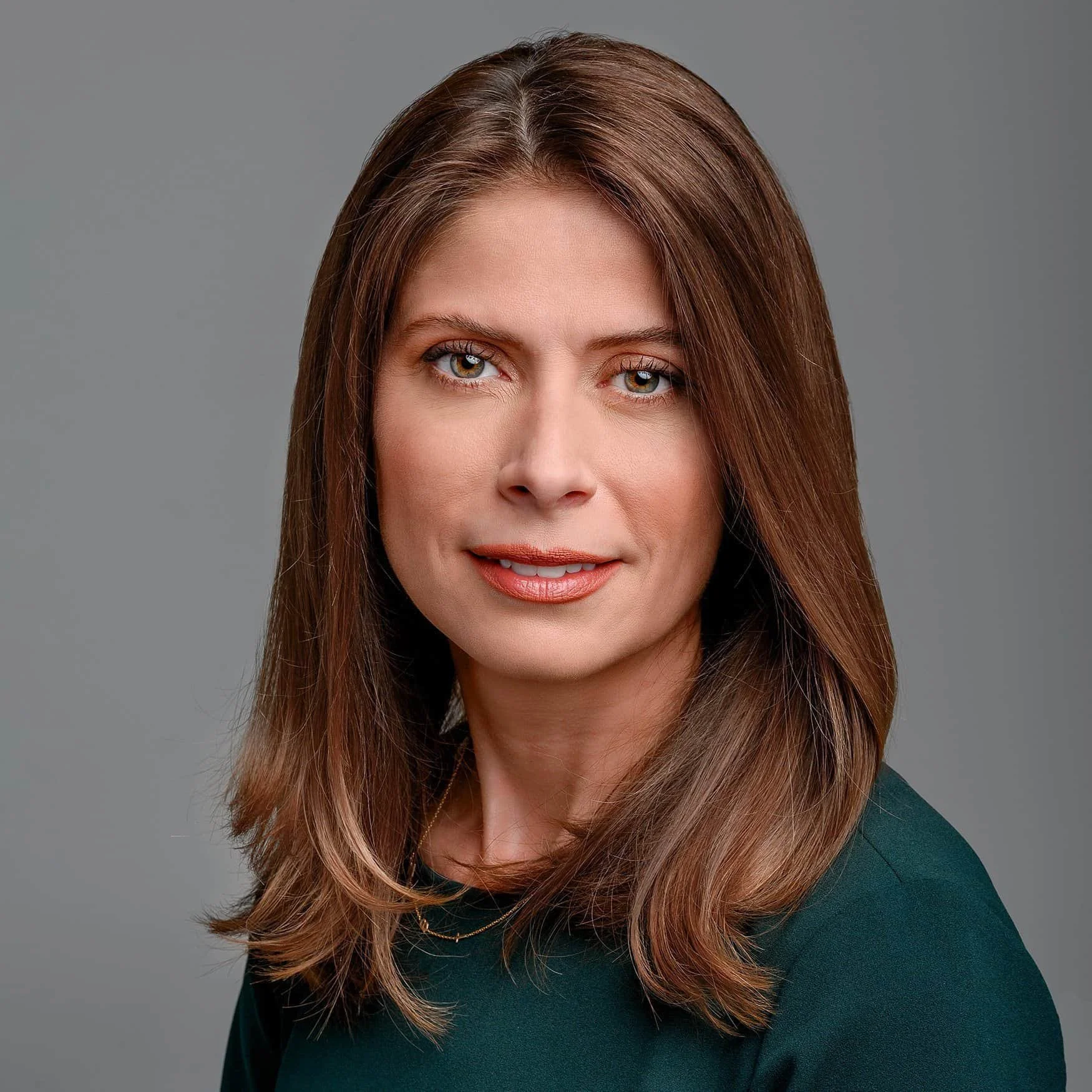 Headshot of a woman with shoulder-length brown hair, wearing a dark green top, smiling slightly against a neutral gray background.