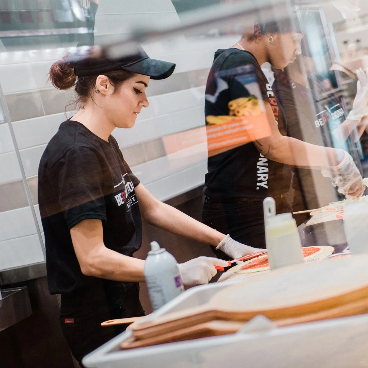 Two women preparing food in a restaurant kitchen, wearing black uniforms and gloves, with one slicing meat and the other working on a different task.