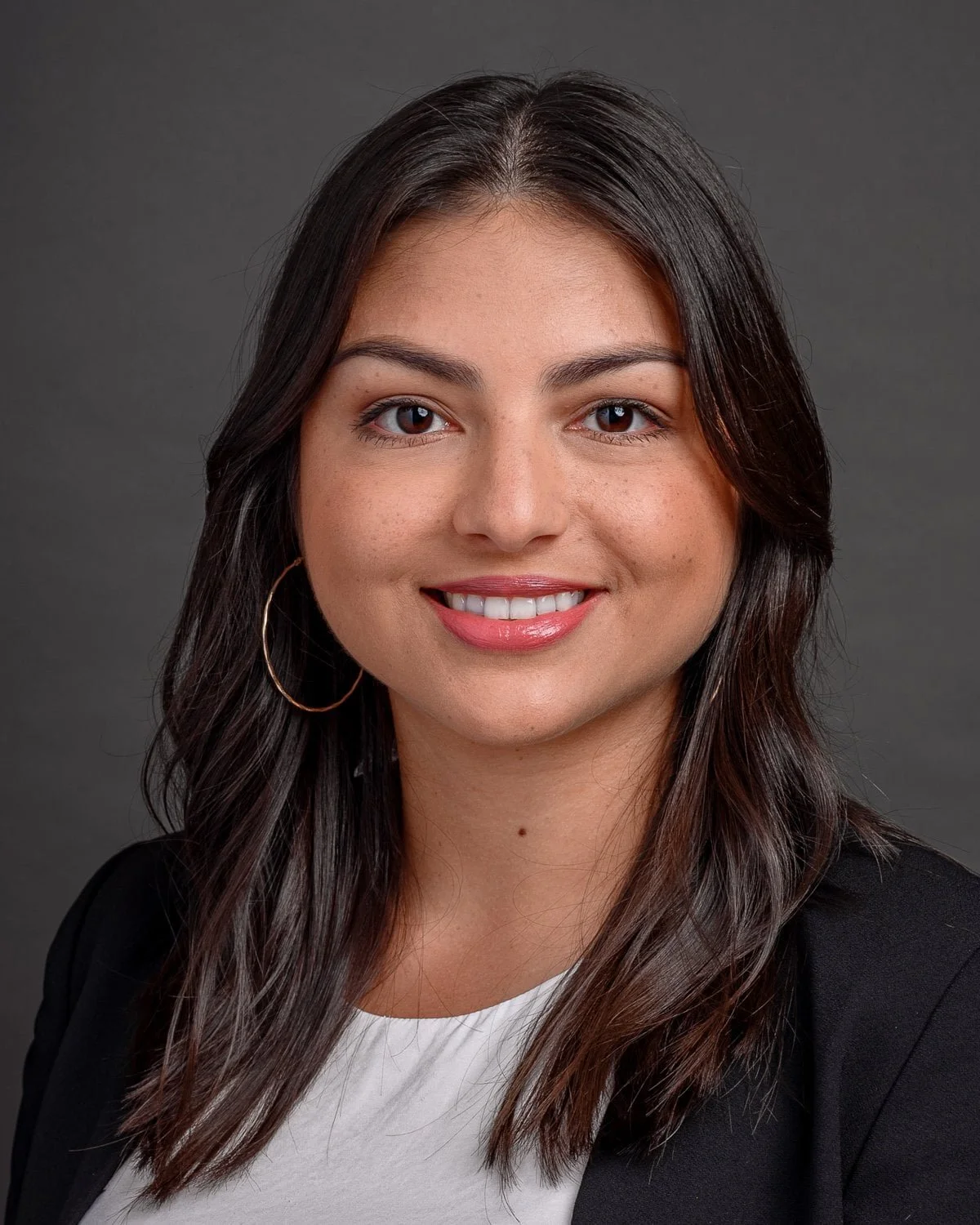 Professional headshot of a woman with long, dark hair, wearing gold hoop earrings, a white top, and a black blazer, smiling against a dark gray background.