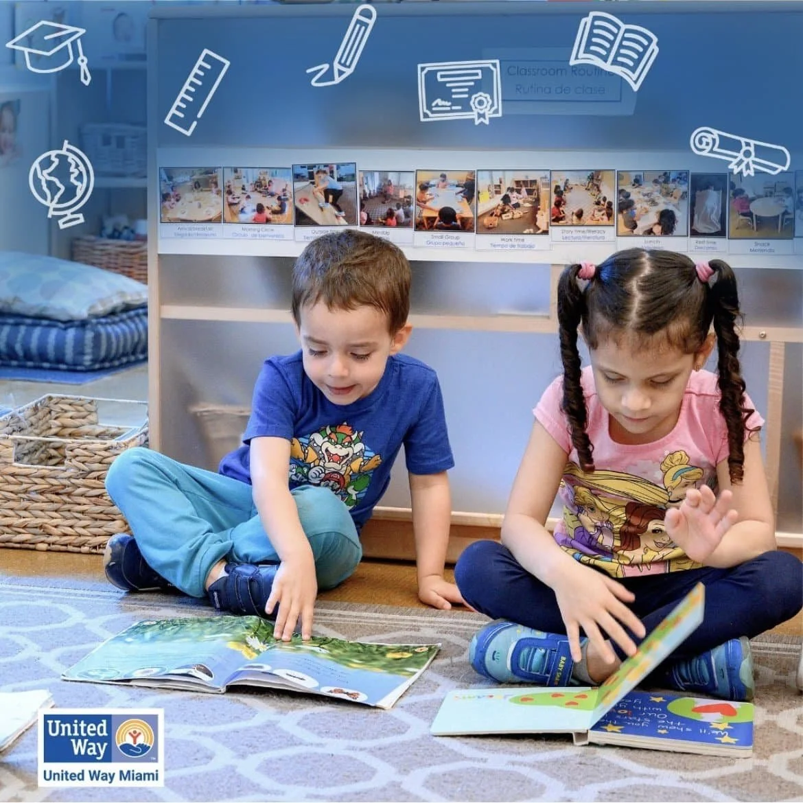 Two children sitting on a carpet, reading picture books in a classroom or library, with educational posters and photos displayed on the wall behind them.
