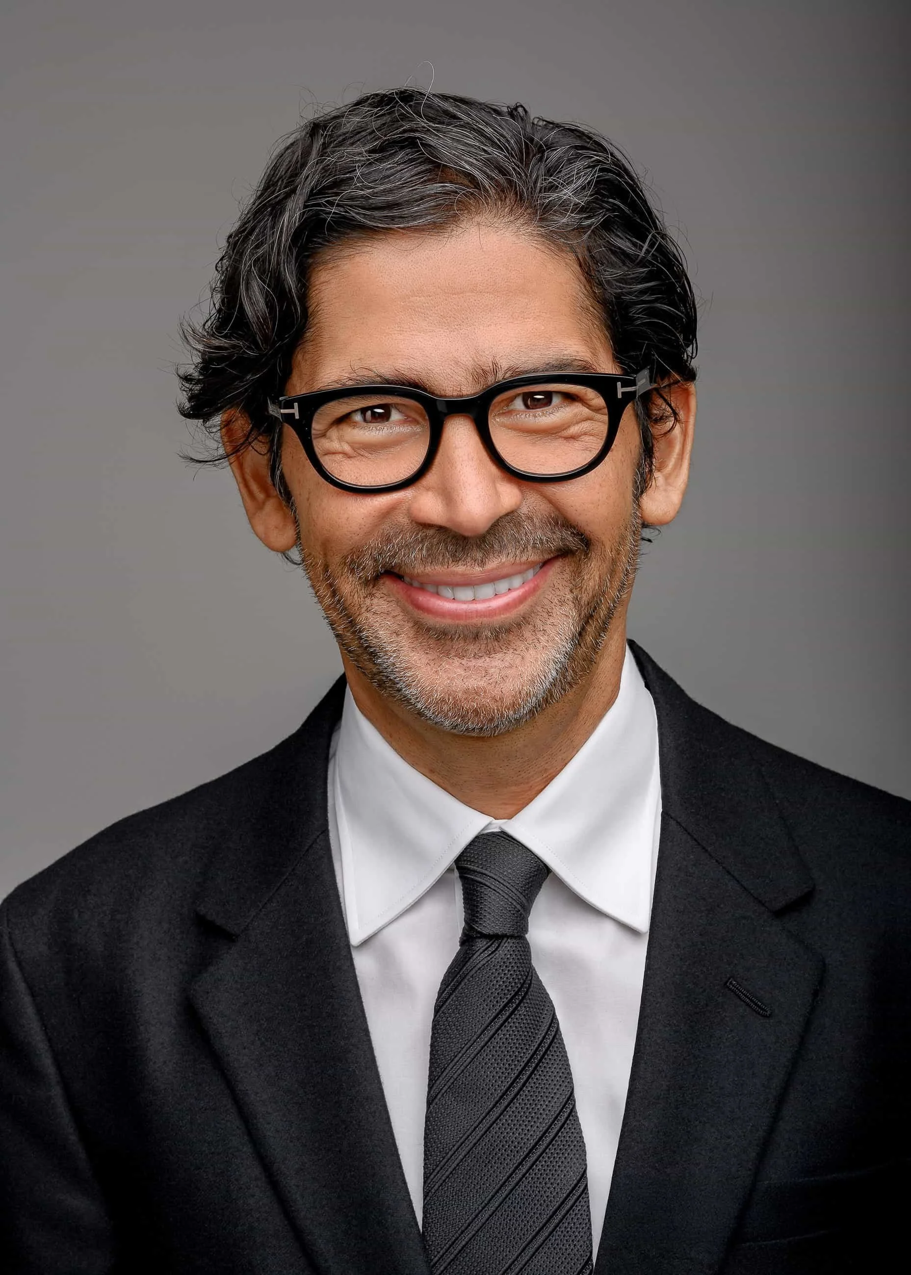 Portrait of a middle-aged man with glasses, dark hair, and a beard, wearing a black suit, white shirt, and a striped black tie, smiling against a gray background.