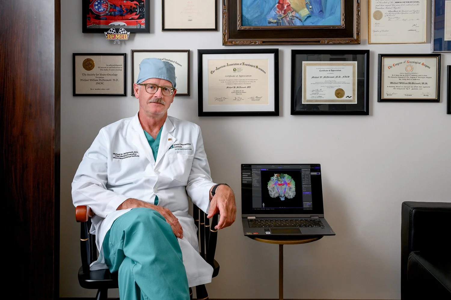 A doctor seated in an office, wearing a white lab coat, green scrubs, and a blue surgical cap. Behind him is a wall decorated with framed certificates and awards. To his right, a laptop displays a colorful brain scan.