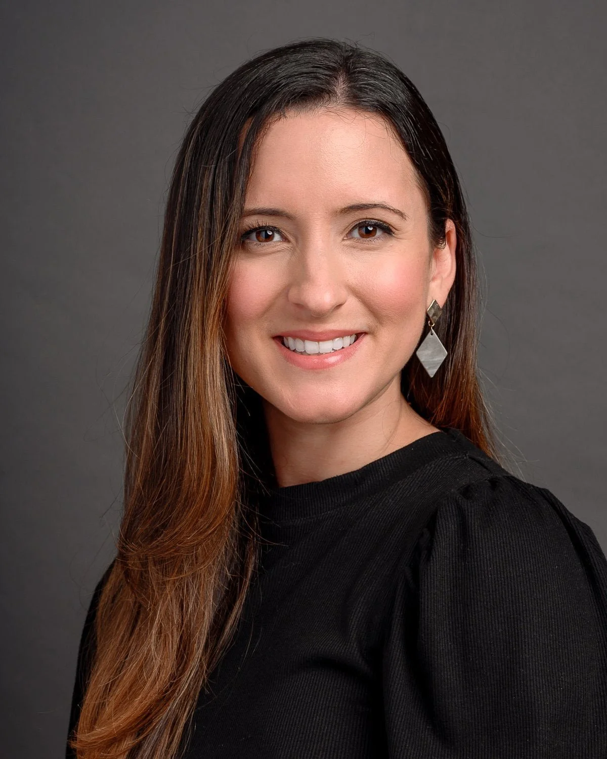 A woman with long brown hair smiling at the camera, wearing a black top and earrings, against a gray background.