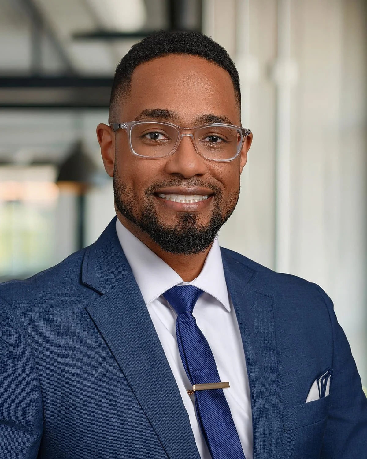 Portrait of a man in a business suit smiling, wearing glasses, with a blurred modern office background.