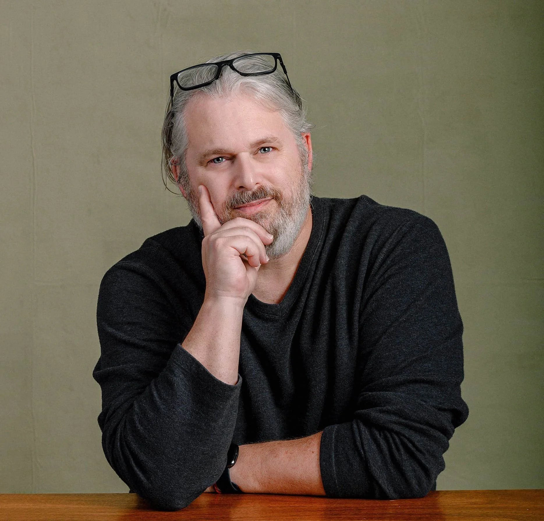 A middle-aged man with gray hair and a beard, wearing glasses on his head and a black long-sleeve shirt, sitting at a wooden table against a plain beige wall, resting his chin on his right hand and smiling gently.