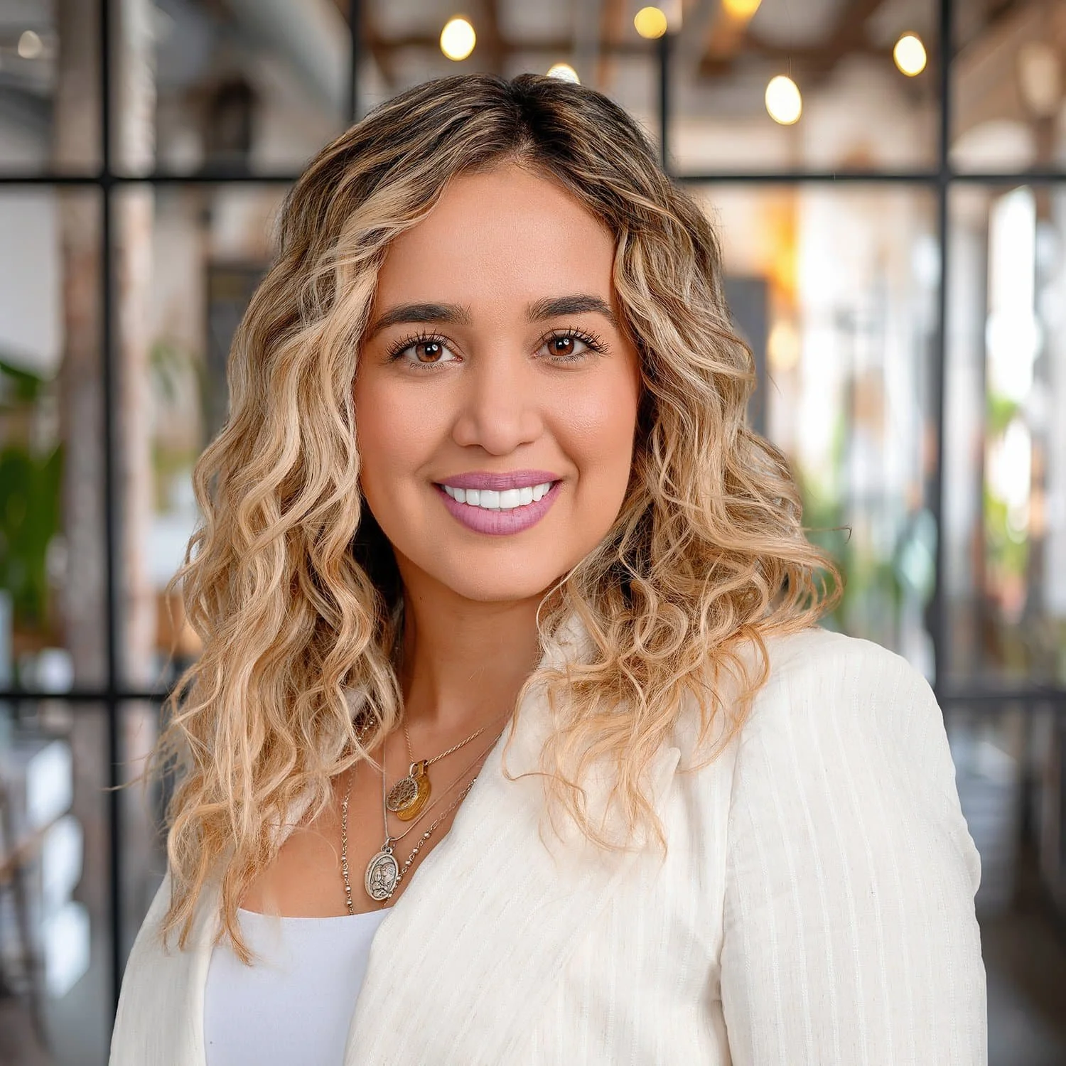 A smiling woman with wavy blonde hair, wearing a white blazer and layered necklaces, in an indoor setting with large window panes and hanging lights in the background.