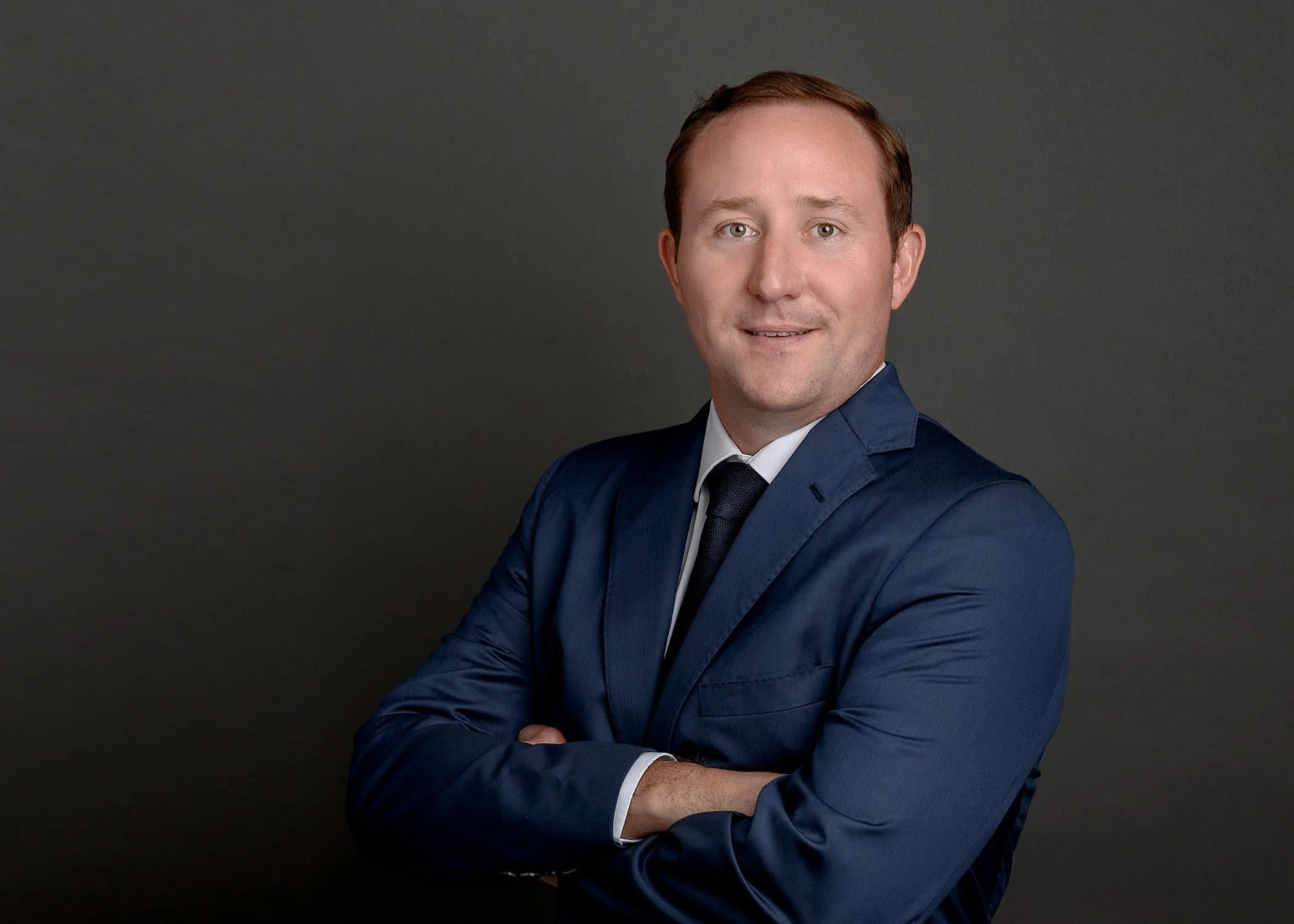 Professional headshot of a man in a blue suit, white shirt, and dark tie, looking at the camera with arms crossed against a dark grey background.