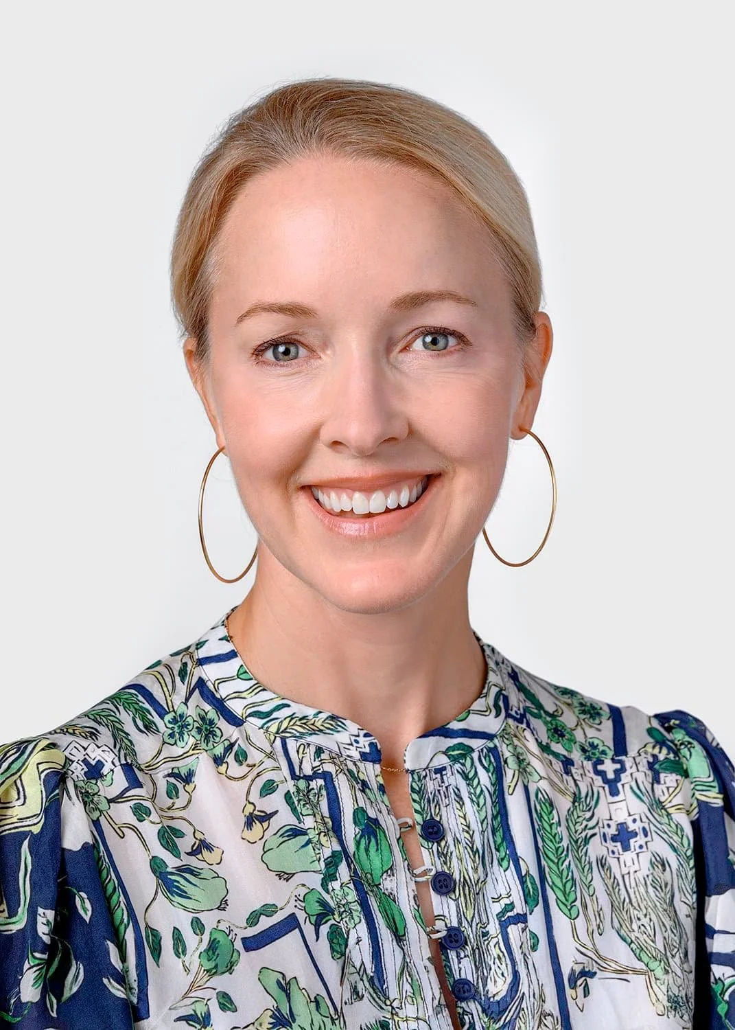 Portrait of a woman with blonde hair, wearing large hoop earrings and a colorful, patterned blouse, smiling against a plain background.