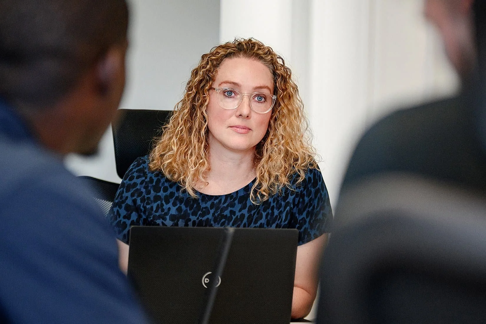 A woman with curly blonde hair and glasses sitting at a desk, facing two men during a meeting.