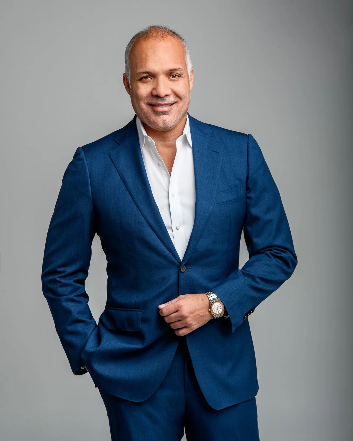 Headshot of a middle-aged man with a bald head, dressed in a blue suit and white shirt, smiling against a plain grey background.