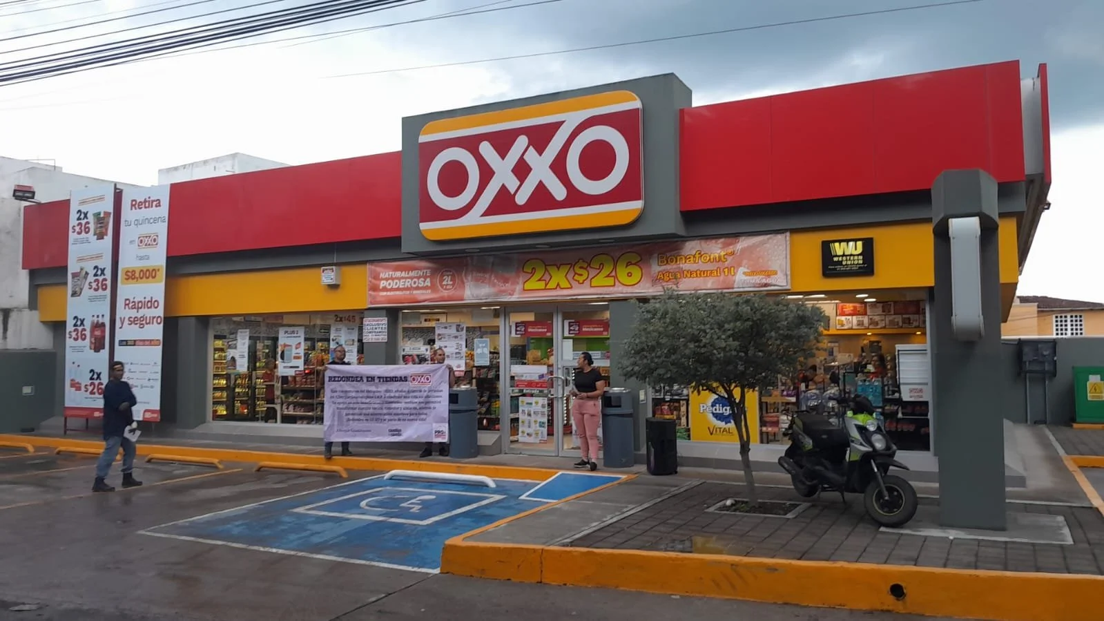 Vista frontal de una tienda OXXO con un letrero grande y señales publicitarias en el exterior, incluyendo promociones y un espacio de estacionamiento para discapacitados.