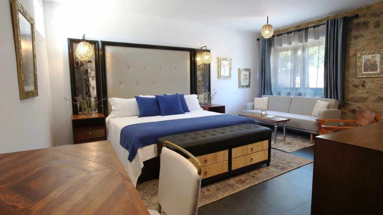 A bedroom with a large bed with white linens and multiple dark blue pillows, a tufted headboard, two nightstands, a sitting area with a white couch, a window with blue curtains, and a stone accent wall.