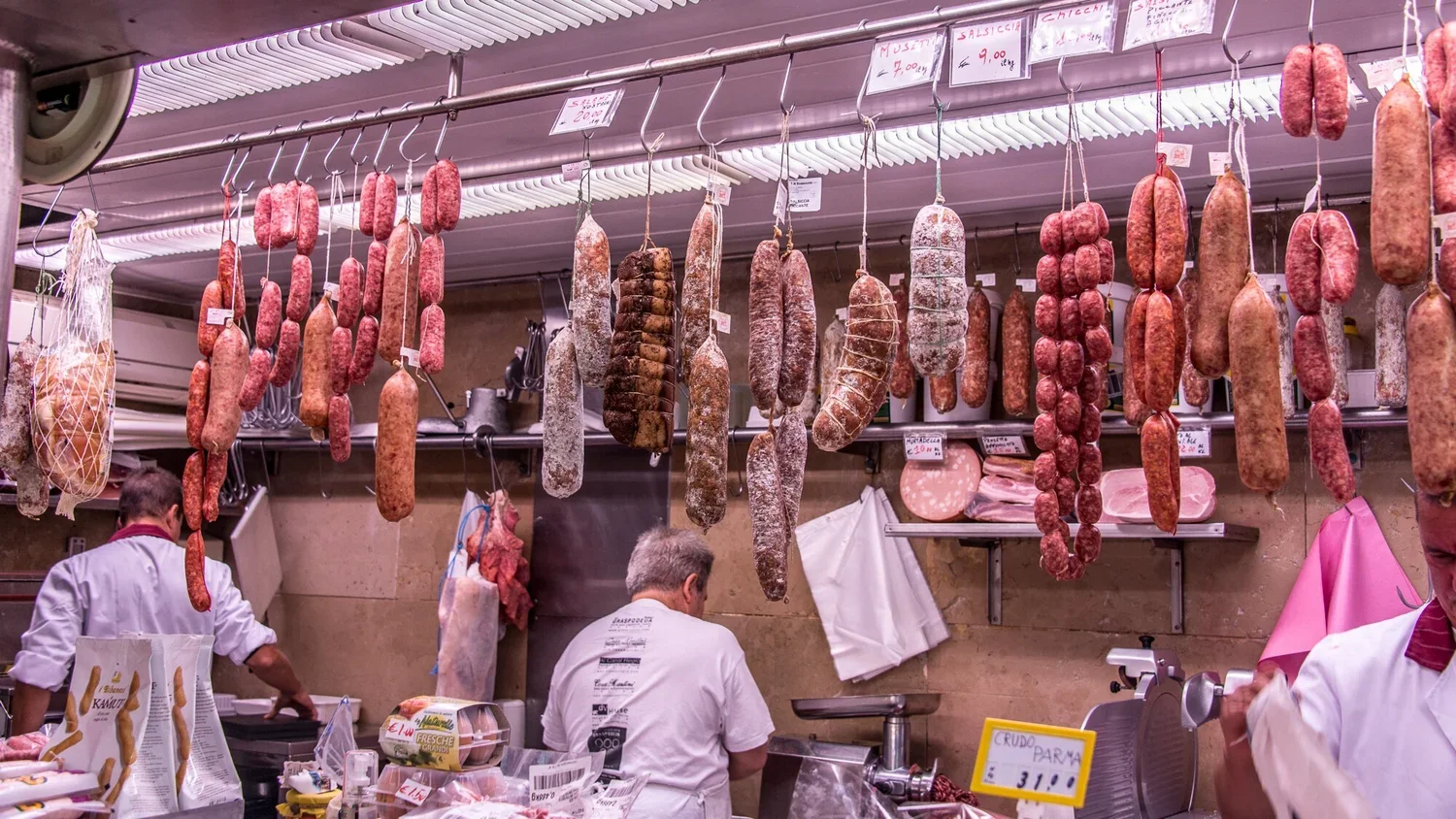 Hanging cured sausages and meats in a butcher shop with workers preparing meat.