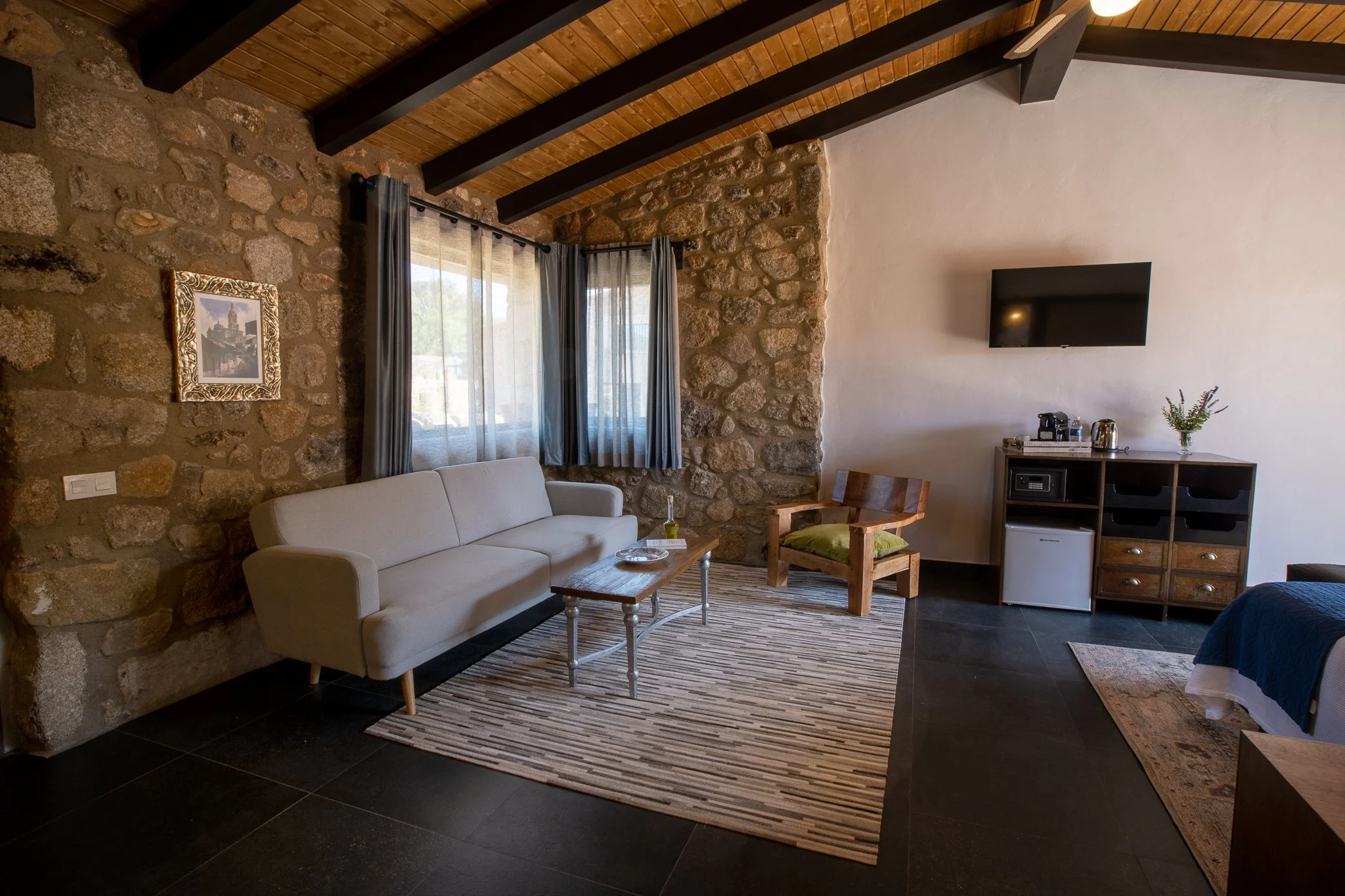 Living room with stone wall, wooden ceiling with dark beams, beige sofa, wooden coffee table, striped rug, window with sheer curtains, armchair with green cushion, wall-mounted TV, small cabinet with coffee maker, kettle, and a plant.