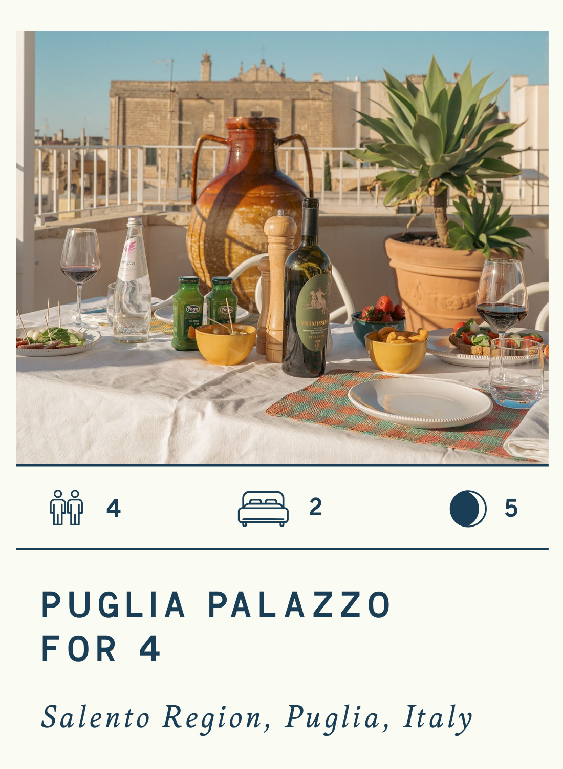 A rooftop dinner table set for four with wine, bowls of food, a bottle of wine, glass of water, plates, and a potted plant, with a cityscape background and warm sunlight, with text describing a luxury palazzo travel package for four Puglia, Italy.
