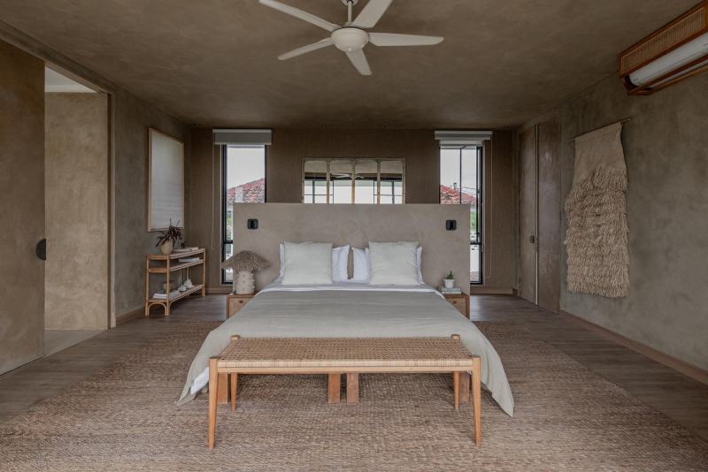 Minimalist bedroom with beige textured walls, a large bed with white pillows, a woven bench at the foot, and small nightstands with decor. Natural light from three windows, ceiling fan, air conditioning, and a woven wall hanging.