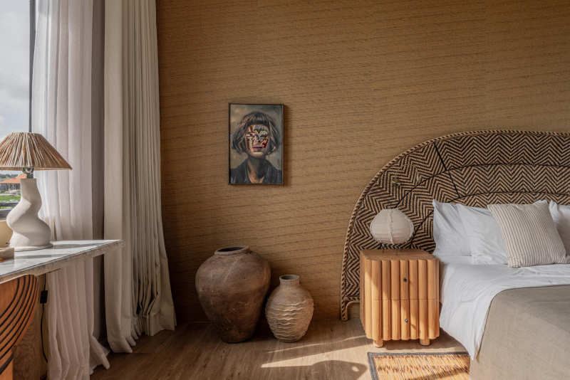A bedroom with a large upholstered headboard, white bedding, and a wooden nightstand with a lamp. There are three decorative ceramic vases on the floor and a framed portrait on the wall. A window with beige curtains is on the left side.