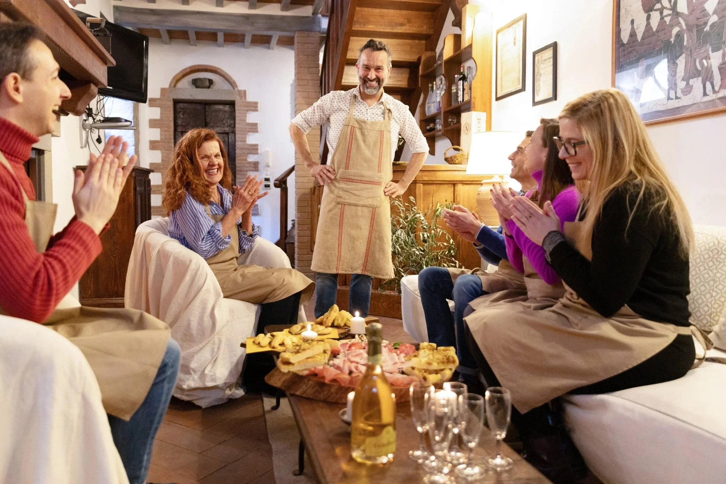 A group of people sitting on couches in a cozy living room, clapping and smiling as a man with a beard, wearing an apron, stands smiling in front of them. A table in front of them holds a bottle of wine, glasses, and a charcuterie board. The room has warm wood accents and framed artwork on the walls.