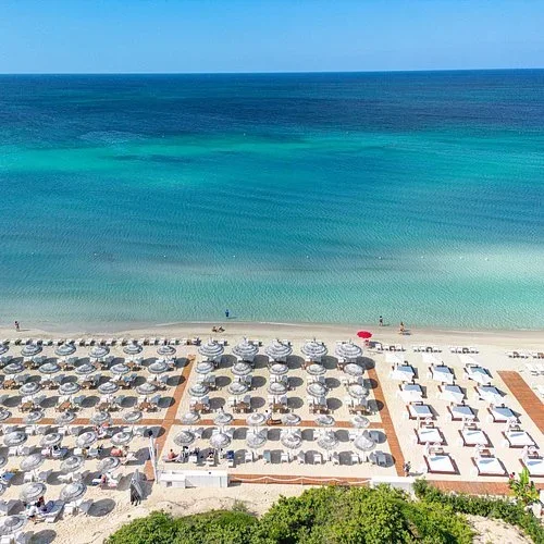 Stone walkway with rusted metal railings leading to an old stone building on a hilltop, overlooking a green landscape and distant cityscape under a blue sky.