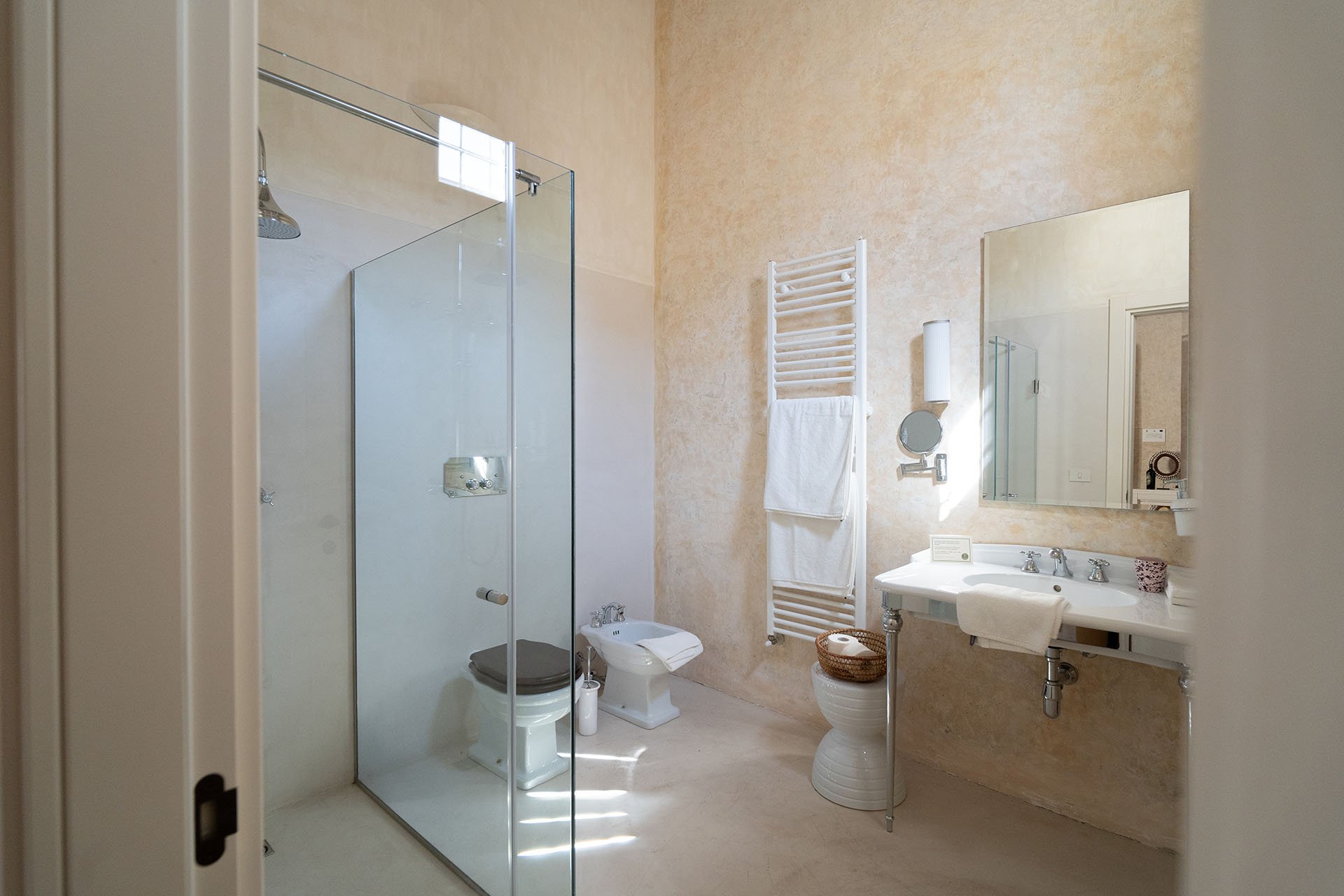 Modern bathroom with a glass shower enclosure, a toilet, bidet, a white towel warmer with towels, a wall-mounted mirror, a small sink with a white towel, and beige textured walls.