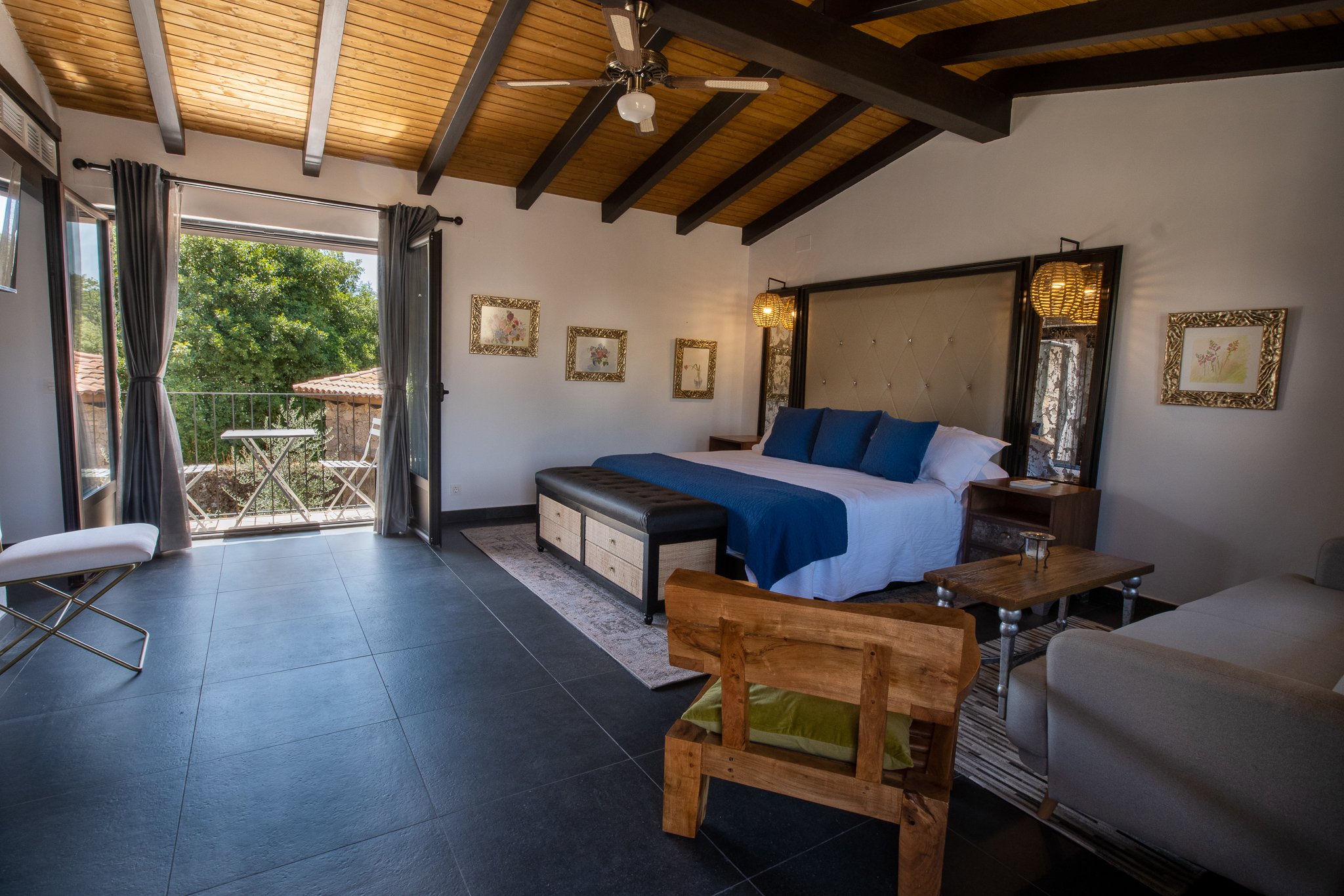 Interior of a bedroom with a large bed with blue pillows and bedding, a wooden bench at the foot, a small wooden side table, and a balcony door with a view of trees outside.