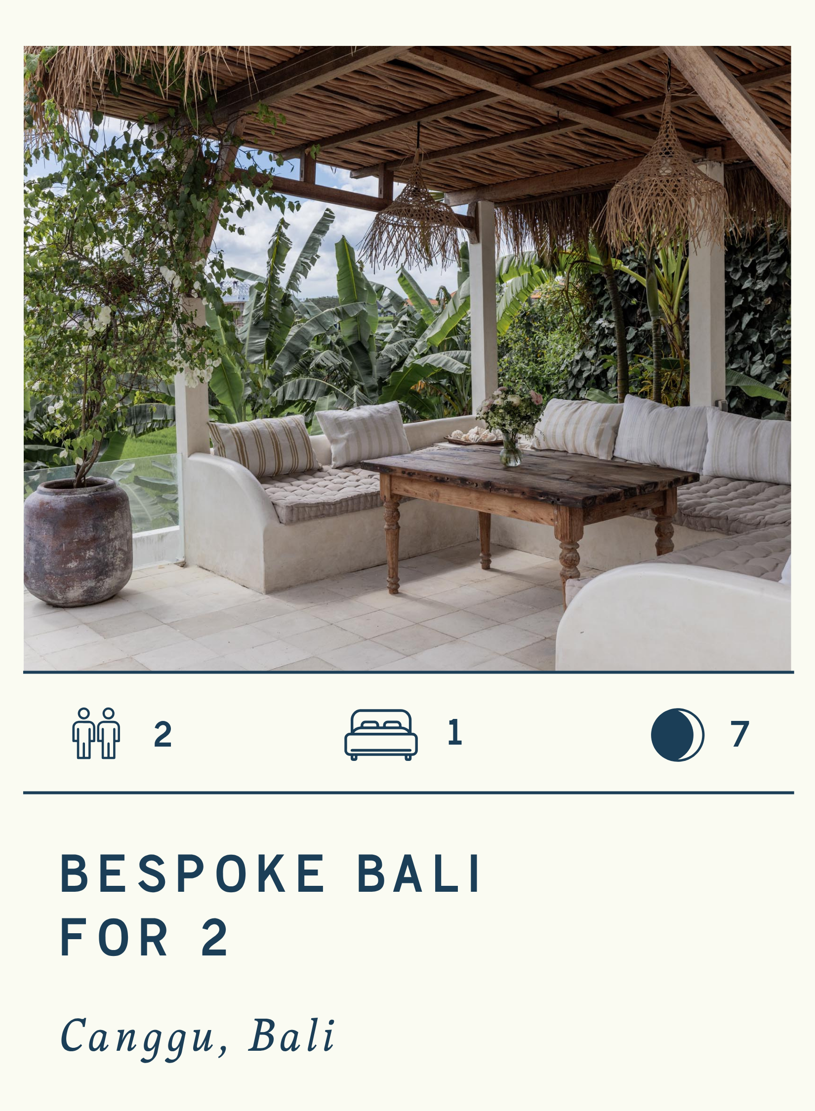 Outdoor seating area with a wooden table, built-in cushioned benches, and pillows in a tropical setting with lush green plants and banana trees in the background.
