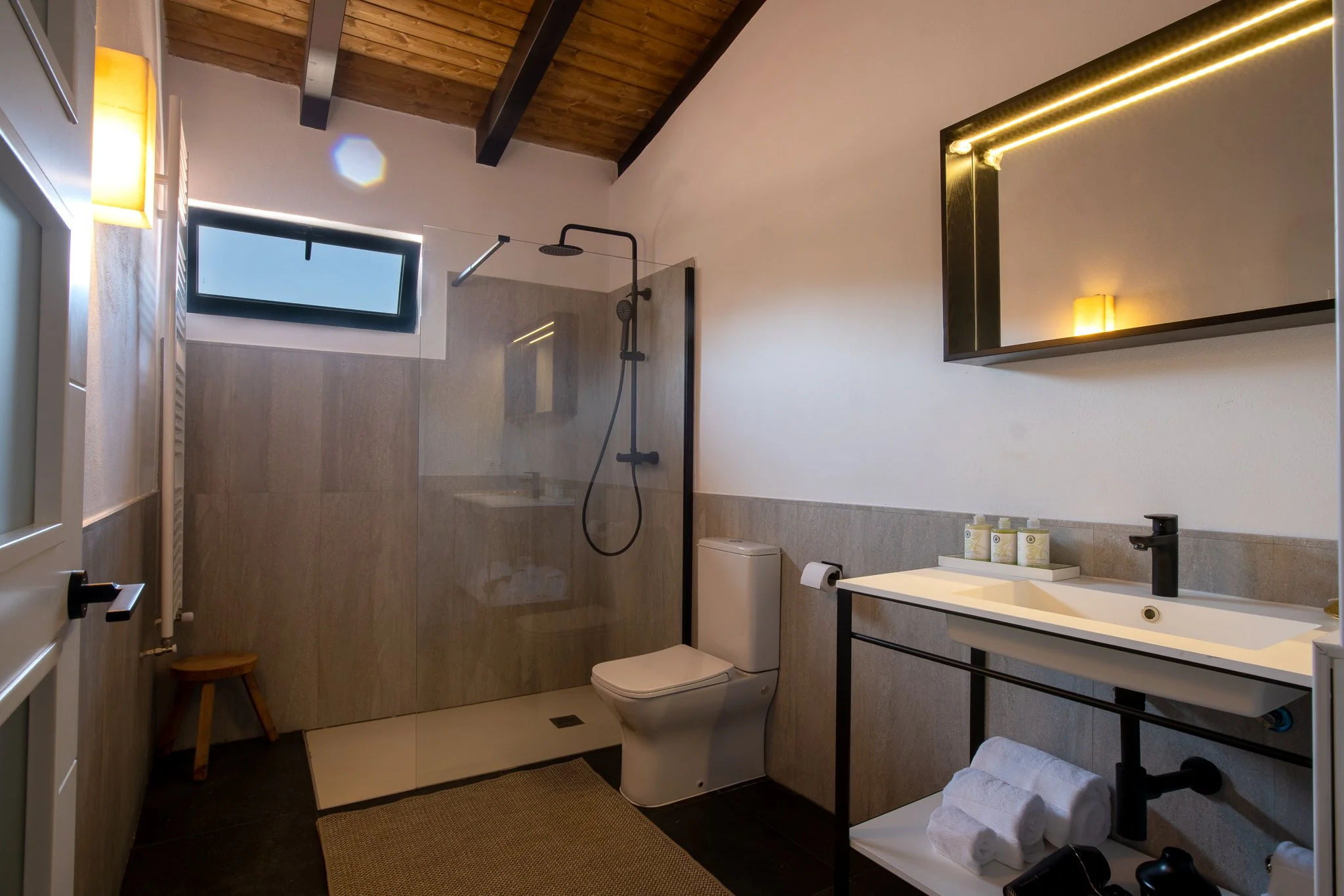 Modern bathroom with a shower area, toilet, sink, mirror, small window, and wooden ceiling beams.