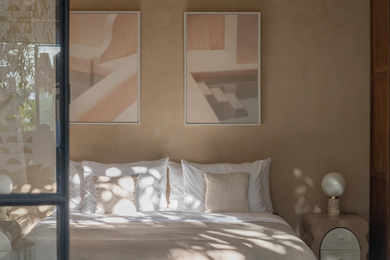 A cozy bedroom with a bed decorated with white pillows and a beige cushion. Two framed abstract art prints hang above the bed. A small beige nightstand with a spherical lamp is on the right side. Natural light filters through a window with blinds and plants outside the window cast shadows on the bed and wall.