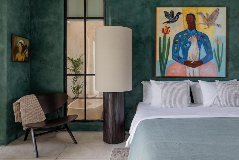 Hotel bedroom with green textured walls, white bed with pillows, large lamp, black chair with beige throw, and colorful artwork of a person with birds and flowers on the wall.