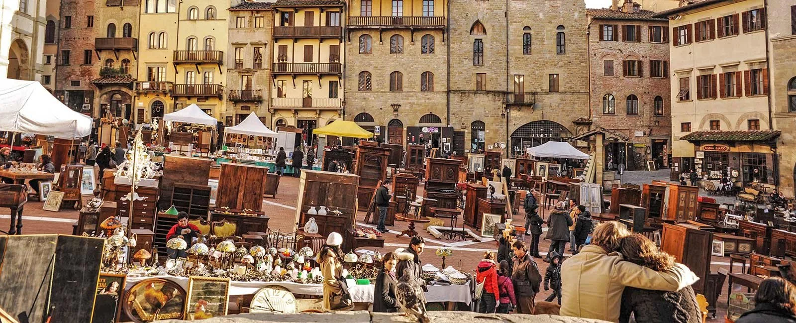 An outdoor flea market in a European city square with vendors and shoppers browsing antique furniture, lamps, and decorative items against historic buildings.