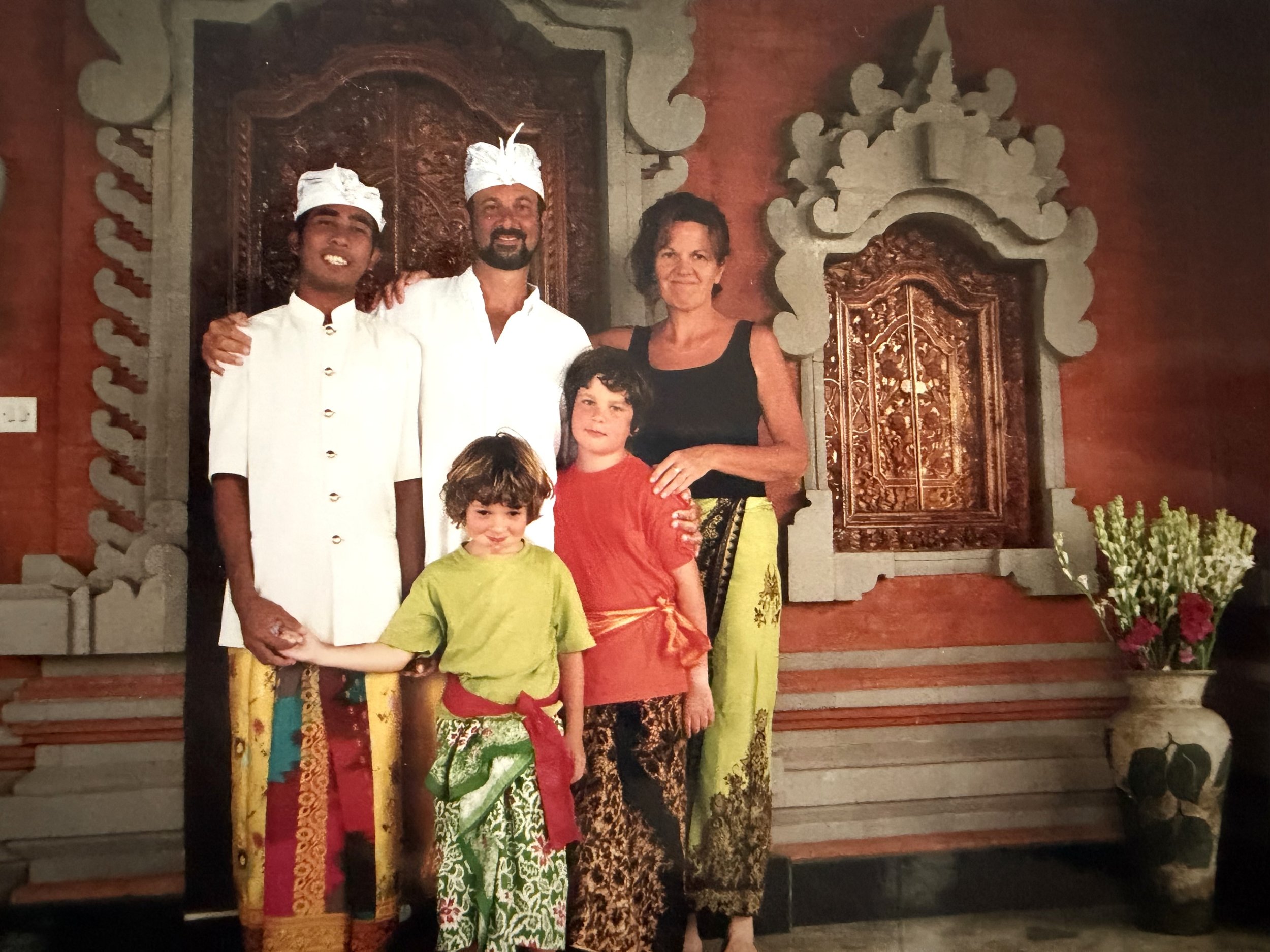 A group of five people standing together indoors, with traditional Balinese decor and flowers in the background. The group includes two young boys, a woman, and two men dressed in traditional Balinese attire.