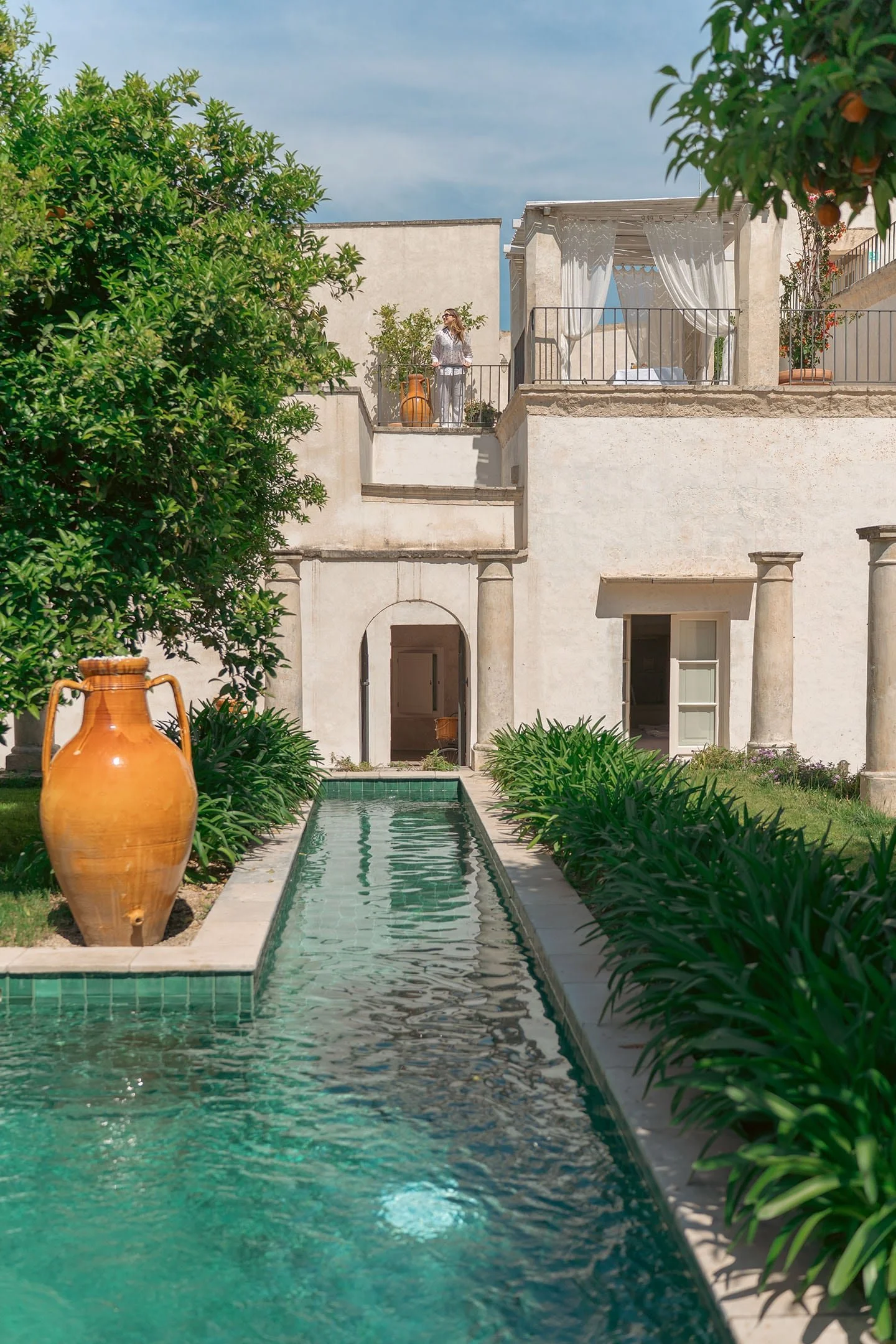 A luxurious outdoor courtyard with a narrow rectangular swimming pool, green plants along its edge, large terracotta vases, a white building with columns, and a woman standing on a balcony under a canopy.
