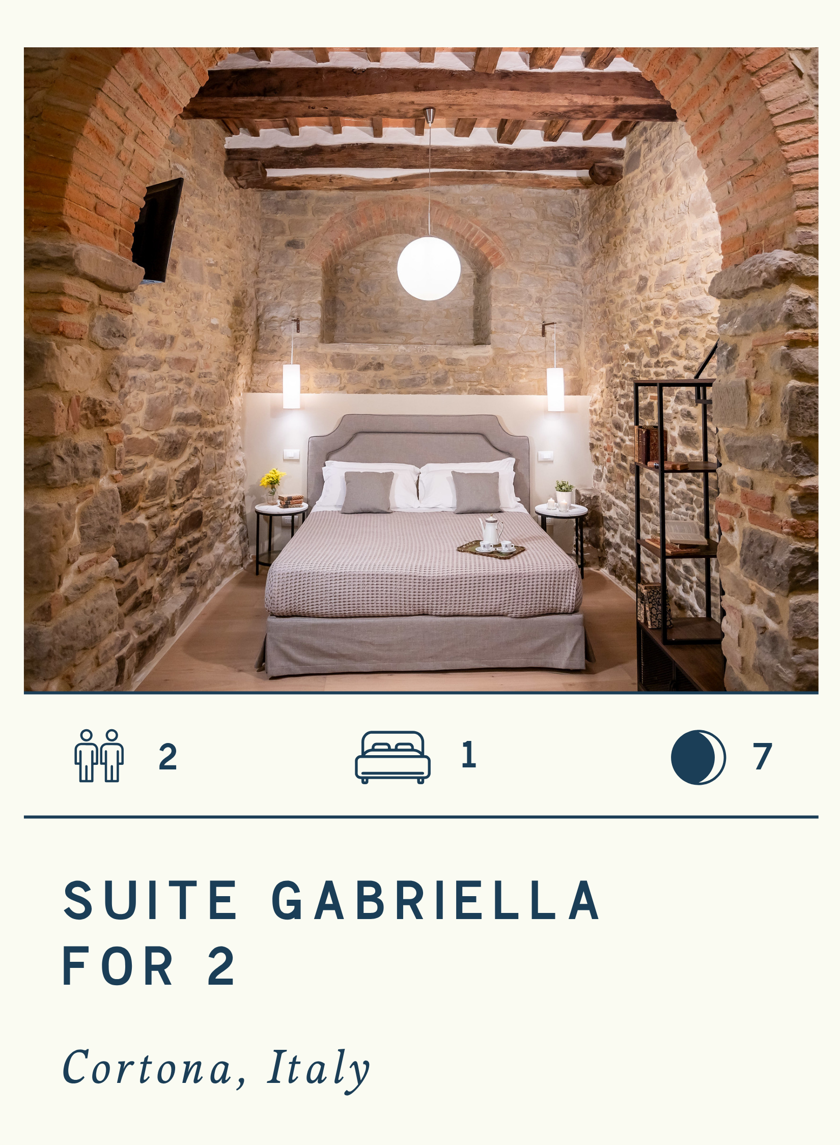 A rustic bedroom in Cortona, Italy with stone walls, wooden beams, and modern lighting. It features a double bed with a gray headboard, white and gray pillows, bedside tables with flowers and a tray, and a black shelving unit.