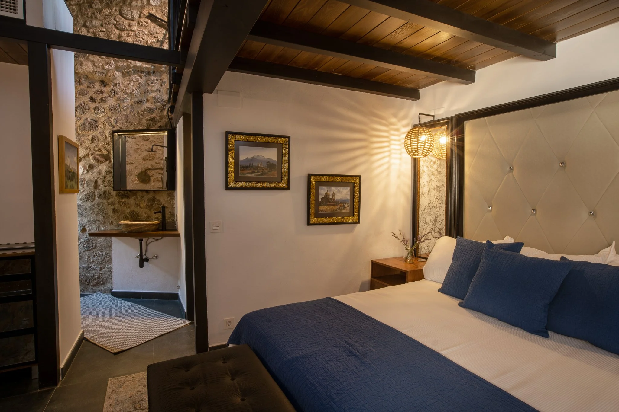 A cozy bedroom with a large bed, navy blue bedding and pillows, a bedside table with a flower vase, framed landscape paintings on the wall, and warm lighting from a woven pendant lamp. There is a stone wall and wooden ceiling.