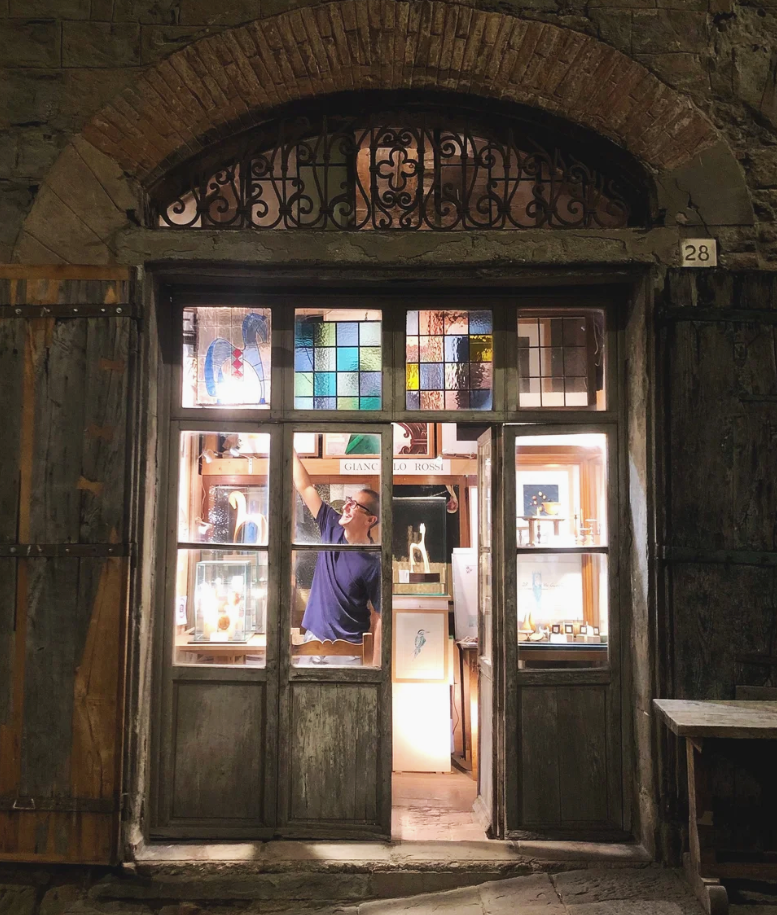 A woman inside a rustic shop, seen through a glass window with patterned stained glass at the top, arranging jewelry or decorative items. The shop has wooden shelves and display cases with various items, with a focus on the woman reaching up.