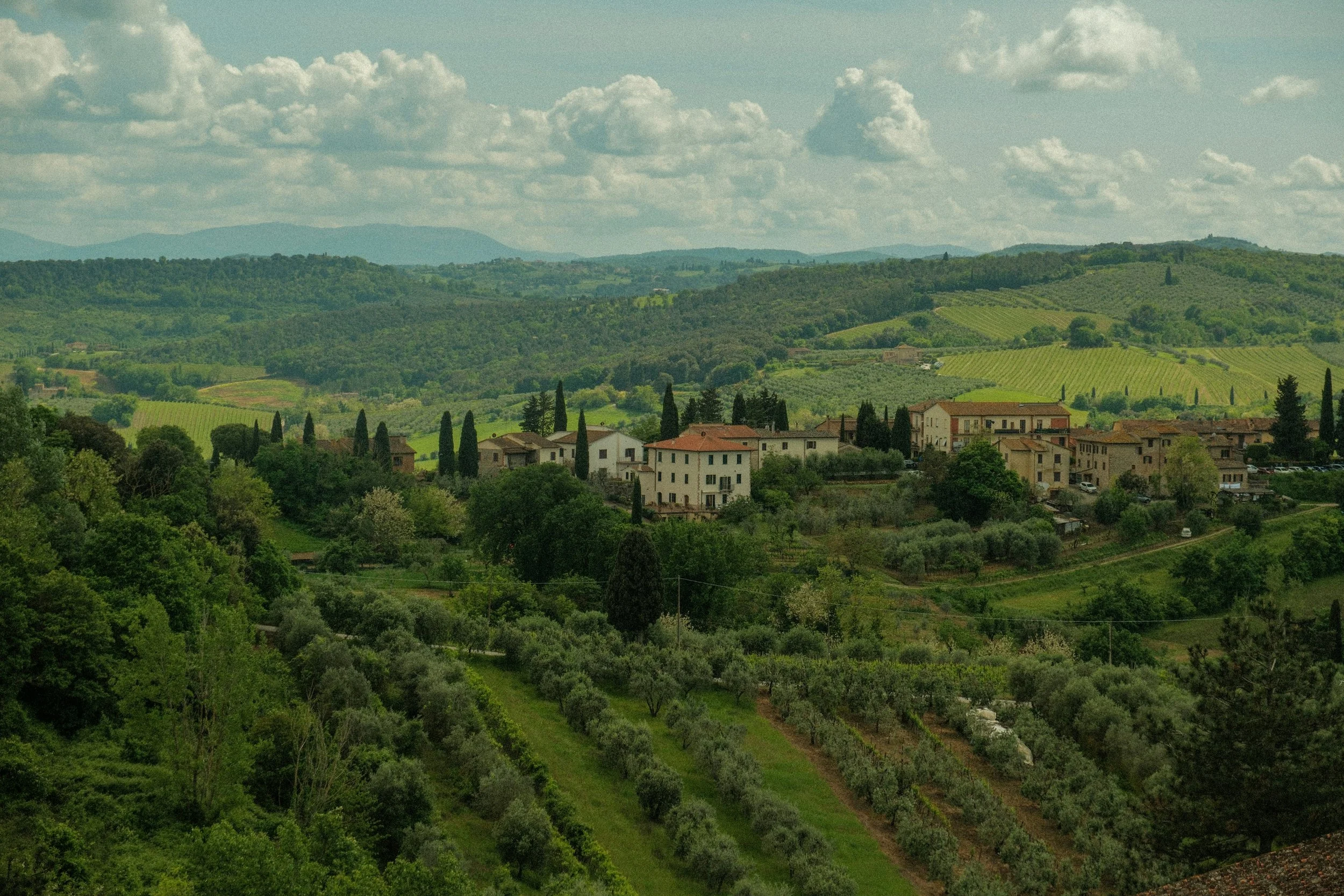 Scenic view of rolling green hills, vineyards, and a small village with multiple buildings and cypress trees under a partly cloudy sky in Tuscany, Italy.