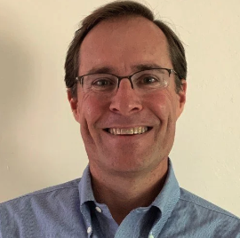A smiling man with glasses in a light blue collared shirt.