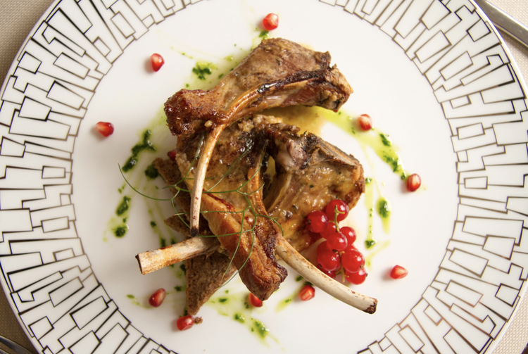 A white plate with a black geometric pattern around the rim, containing cooked lamb chops, garnished with redcurrants, green herbs, and sauce drizzles.