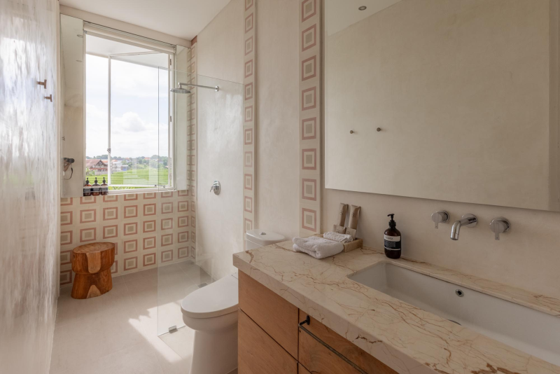 Bathroom with a window, patterned tiles, a wooden stool, a toilet, a marble countertop, a sink, a soap dispenser, and a mirror.