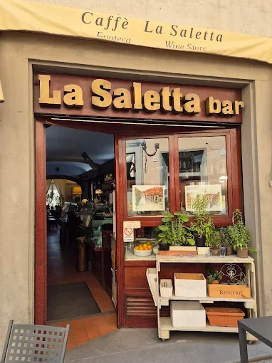 The storefront of La Saletta bar with a yellow awning and large wooden sign, displaying potted plants and a small shelf outside the entrance.
