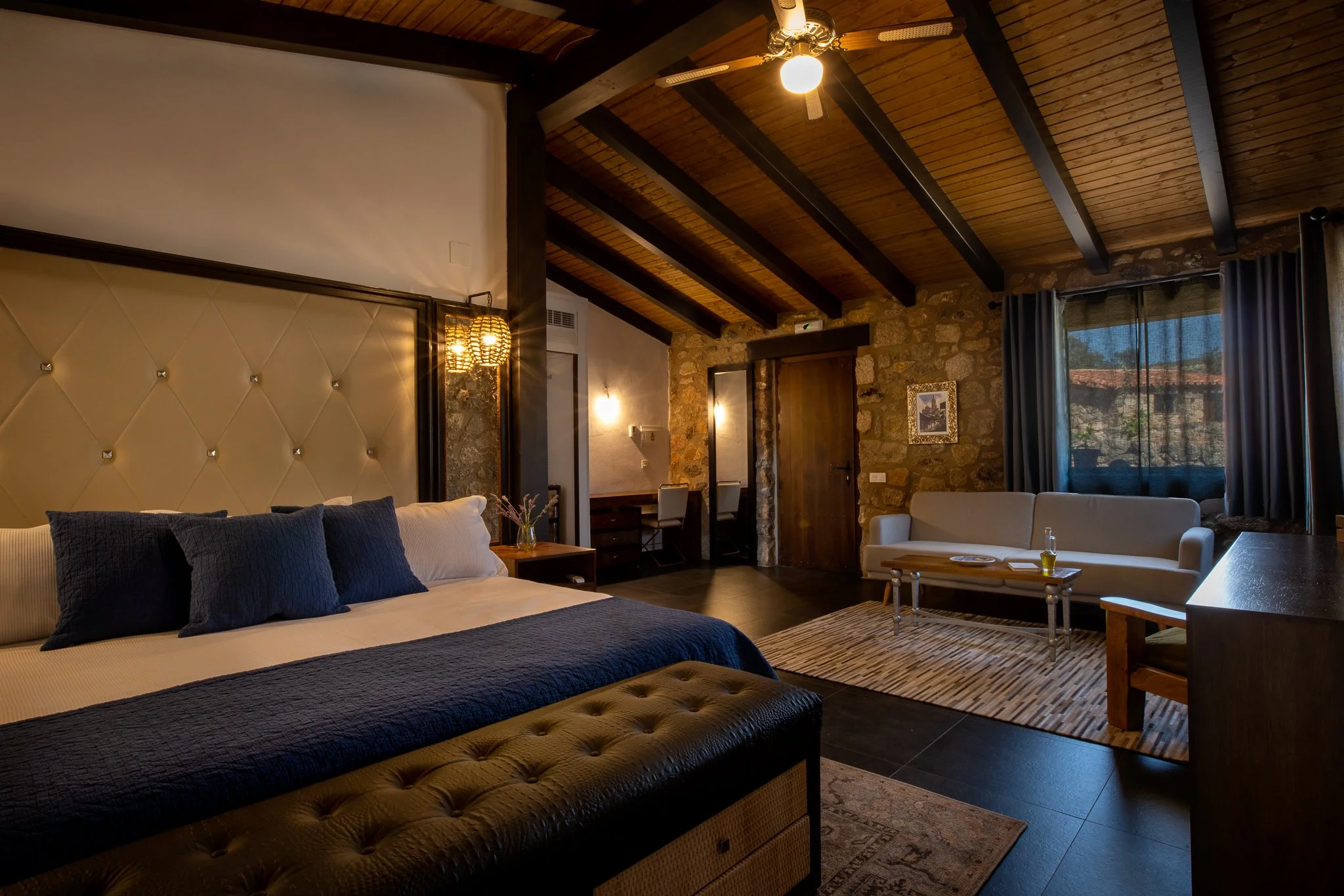 Hotel room interior with a bed, dark blue pillows, beige headboard, small sitting area with a white couch, wooden coffee table, and a window with blue curtains. Exposed wooden beams and stone wall add rustic charm.