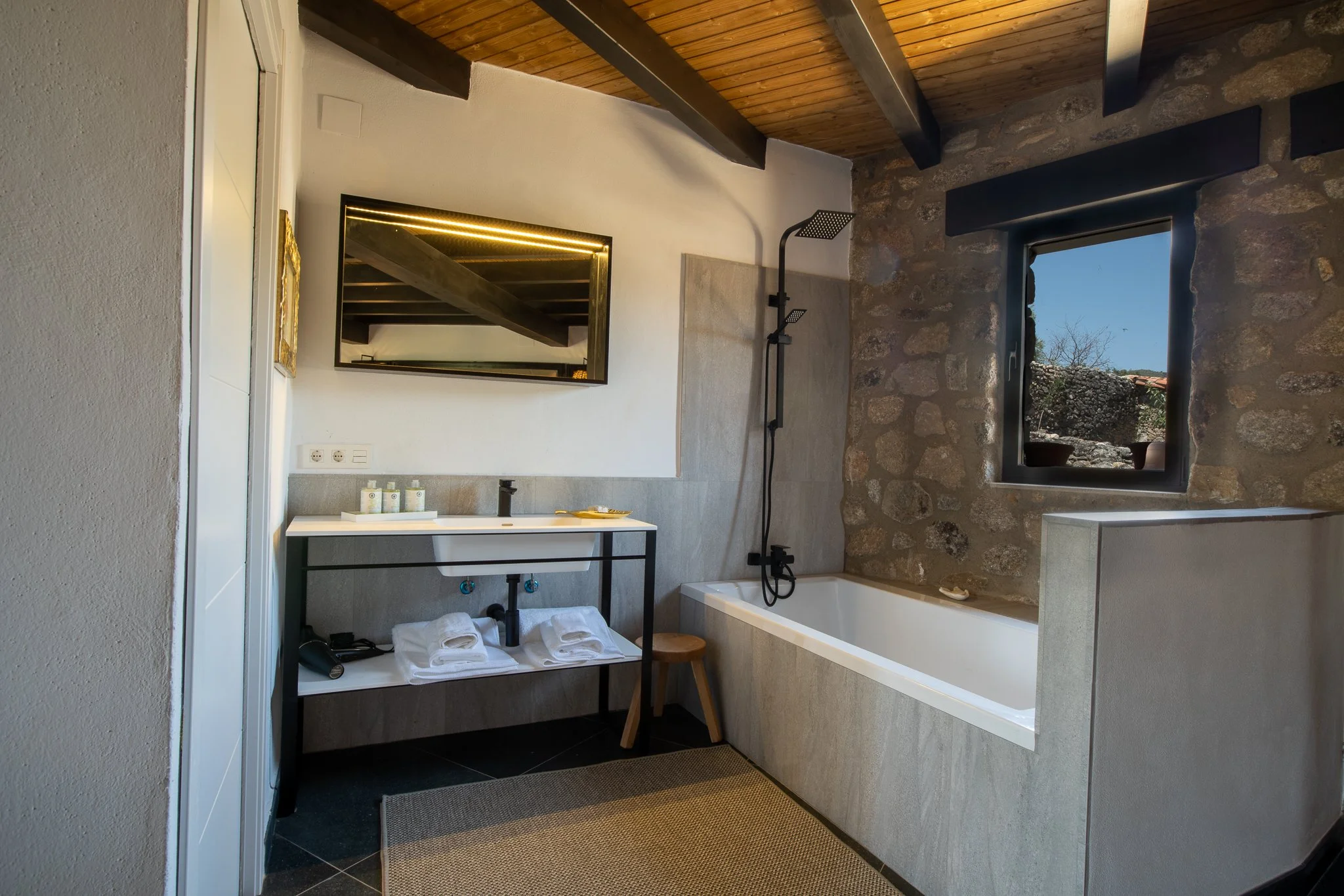 A bathroom with a rustic stone wall, a large window, a bathtub, a black showerhead, a mirror, a sink with toiletries, and towels on a shelf.