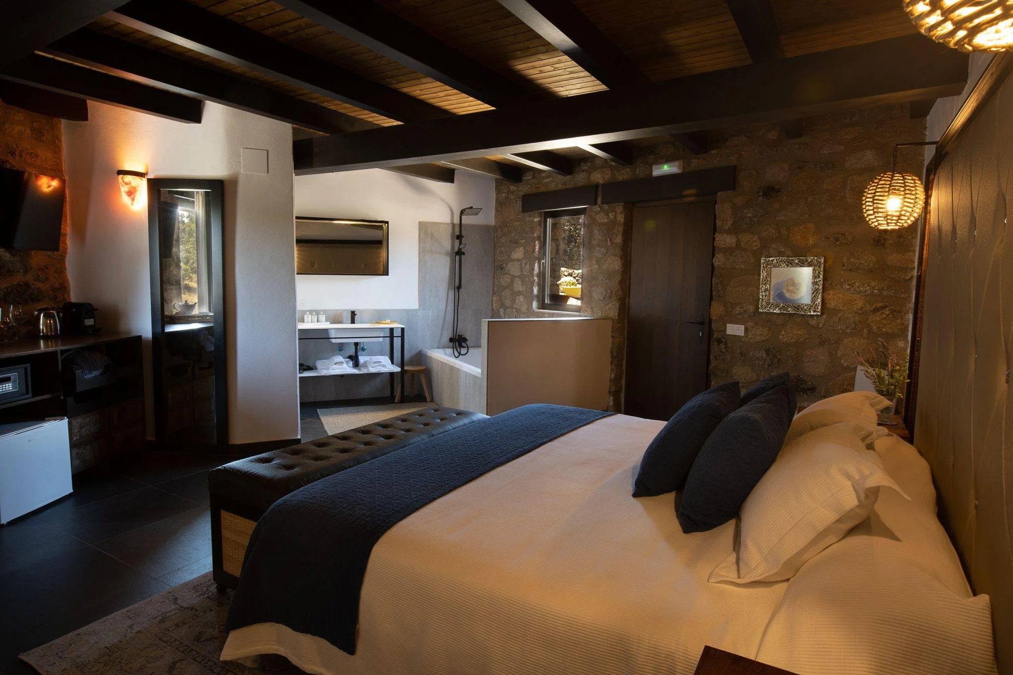 A cozy bedroom with a stone accent wall, a large bed with white linens and black pillows, a tufted ottoman at the foot of the bed, and a small bathroom area with a bathtub, sink, and window. The room has dark exposed beams on the ceiling and warm lighting fixtures.