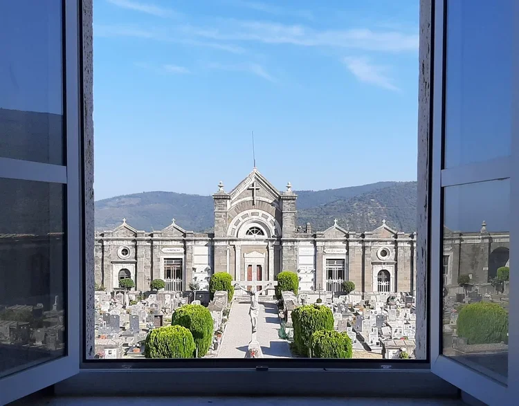 View of a historic cemetery through an open window, with a large chapel in the background and mountains in the distance.