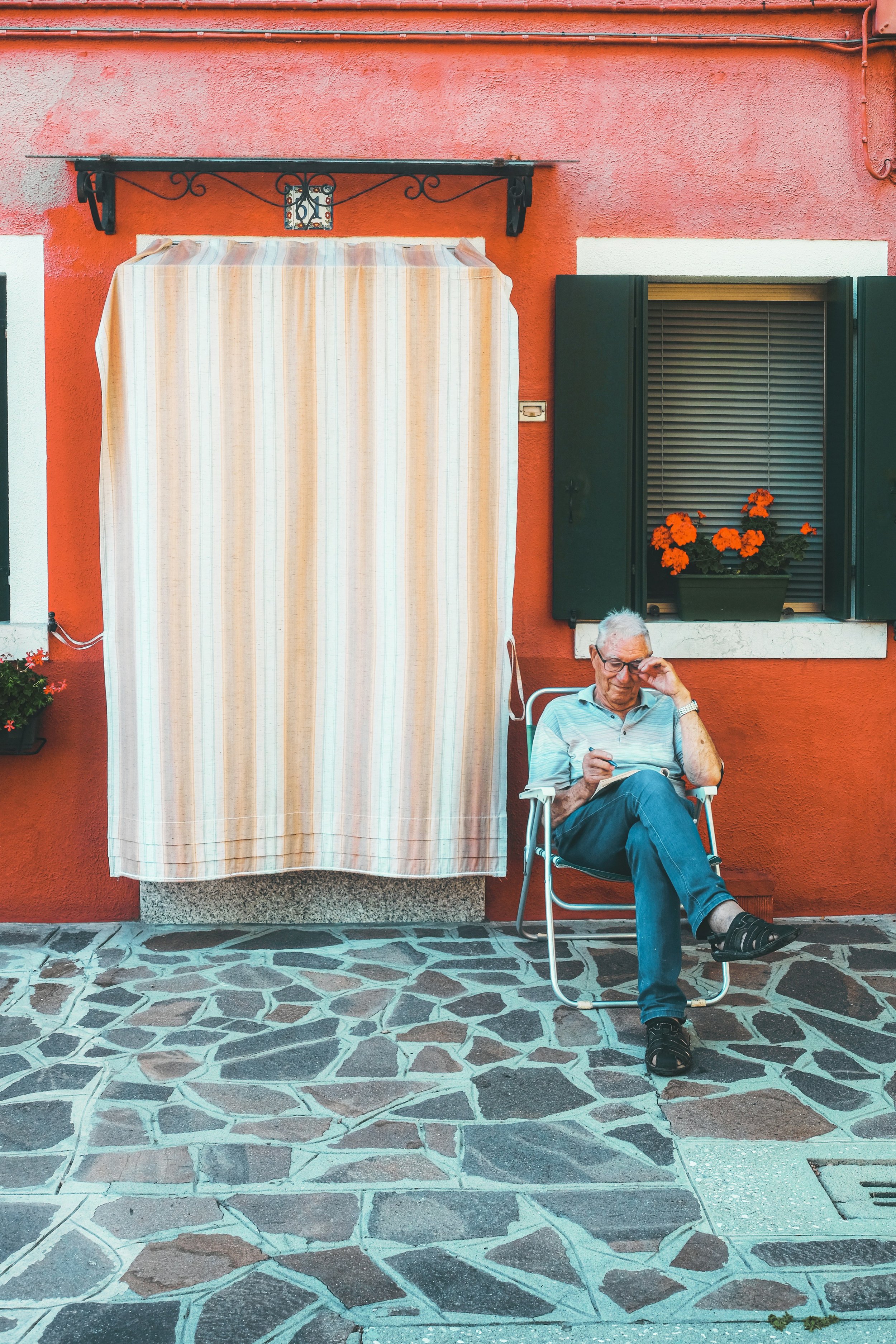 An elderly man sitting on a chair outside a red-orange house, reading a book. There is a window with green shutters and a flower box with orange flowers.