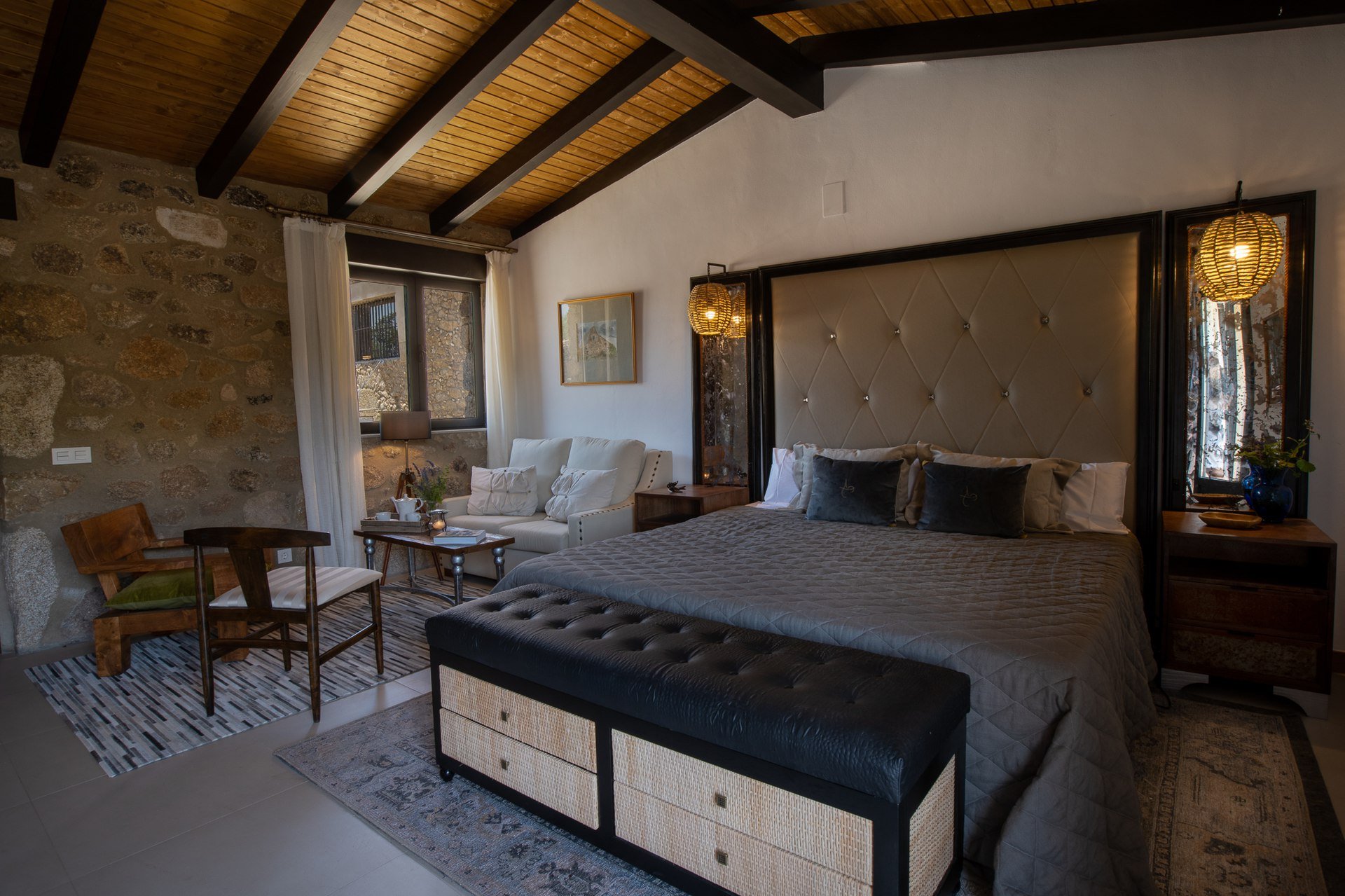 A bedroom with a large bed with a beige padded headboard and dark pillows, two nightstands with lamps, a sitting area with a white sofa, a rustic stone wall, wooden ceiling beams, and a window with curtains.