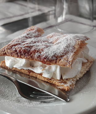 Stone walkway with rusted metal railings leading to an old stone building on a hilltop, overlooking a green landscape and distant cityscape under a blue sky.