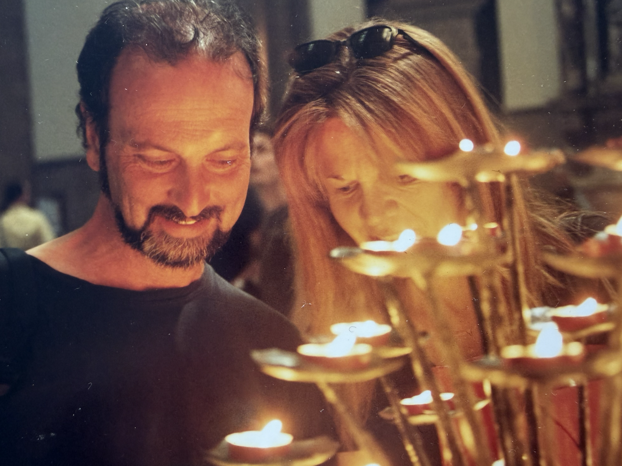 A man and a woman with light skin and hair are smiling and looking at lit candles in a candle holder at night.