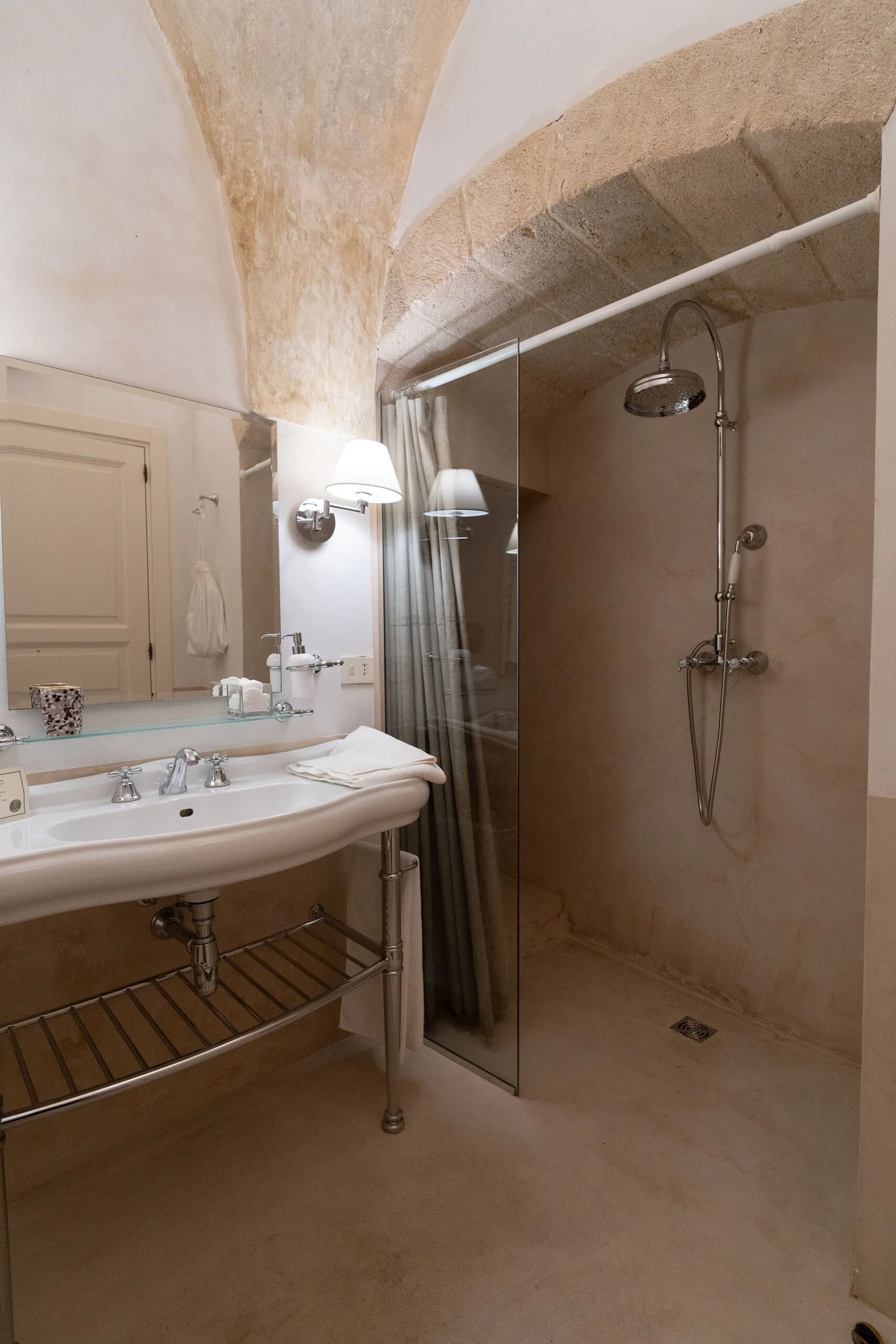 A bathroom with a white porcelain sink, a large mirror, a wall-mounted light fixture, a glass shower partition, and a showerhead on a stone wall. There is also a beige curtain and a wooden shelf under the sink.