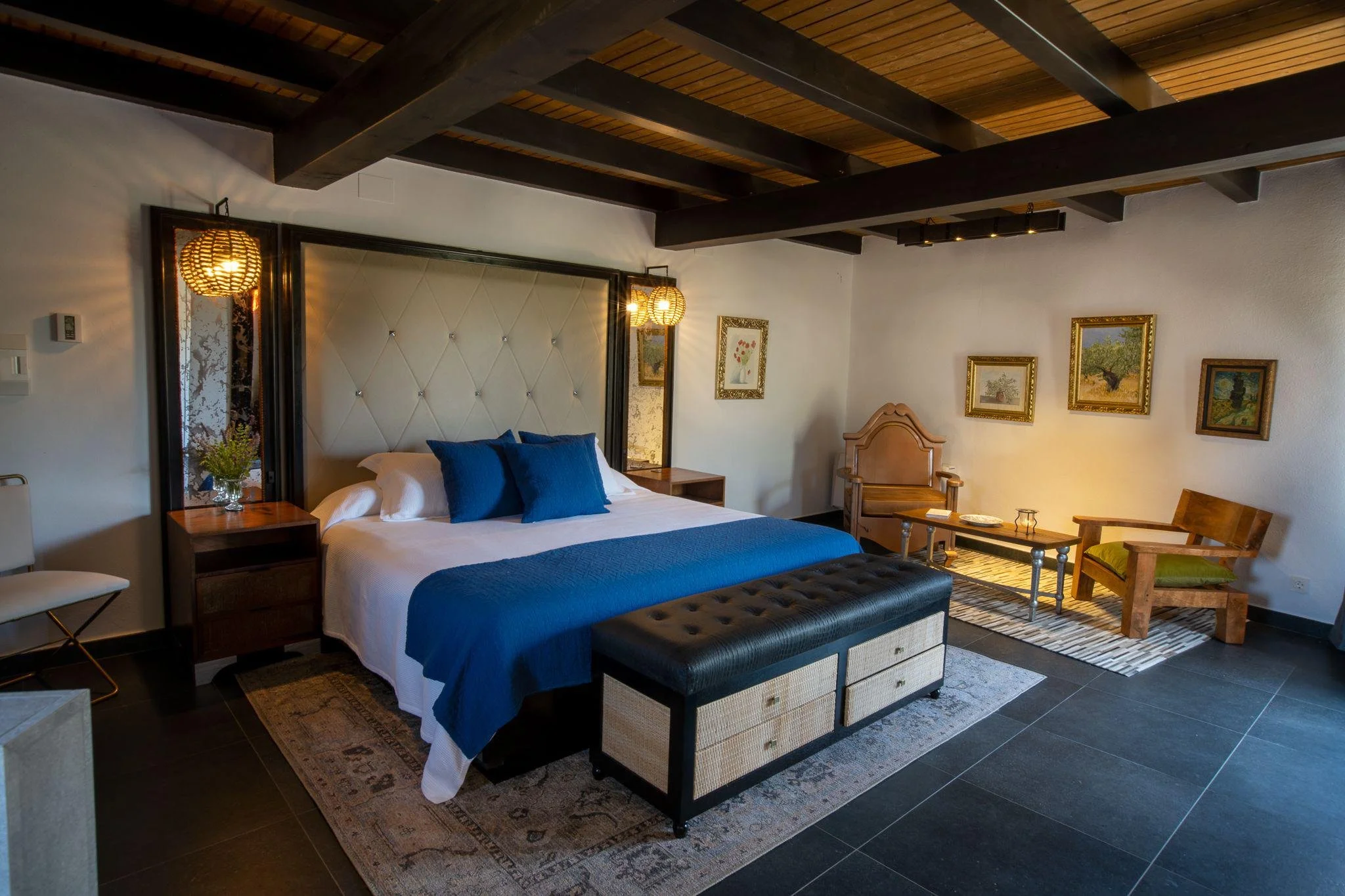 Interior of a bedroom with a large bed with white and blue bedding, a big tufted headboard, side tables, and artwork on the walls. There is a sitting area with chairs and a coffee table, and the ceiling has exposed wooden beams.